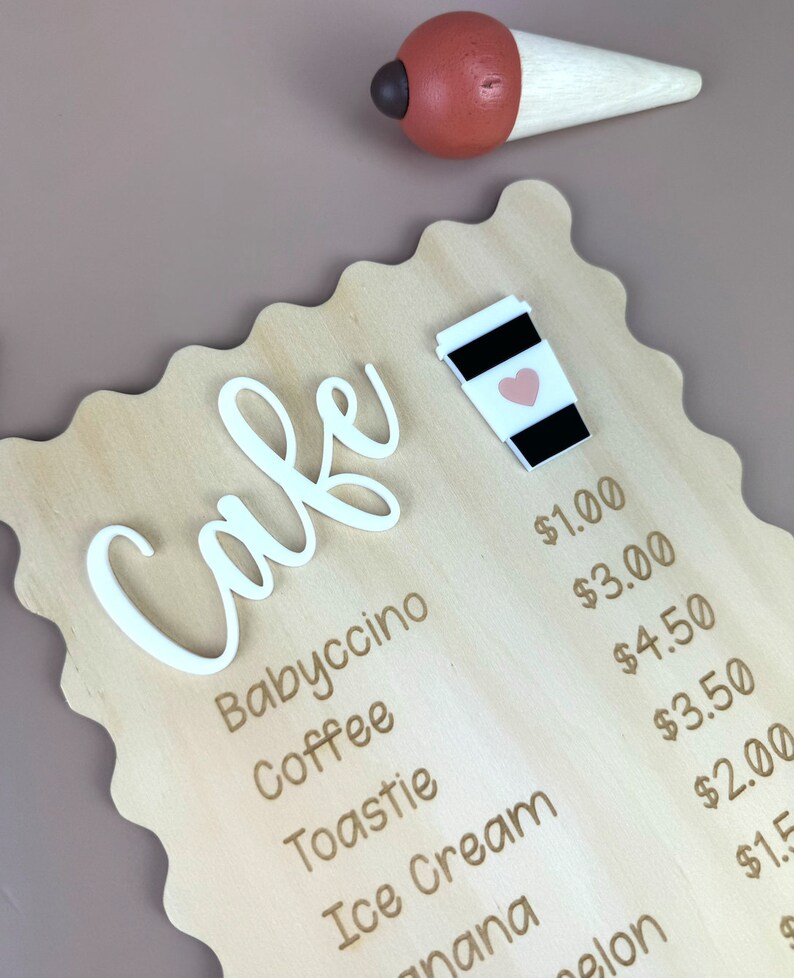 May include: A wooden sign with a white cursive word "Cafe" and a price list for various drinks and desserts. The prices are listed in US dollars. The sign is decorated with a small white coffee cup with a pink heart.