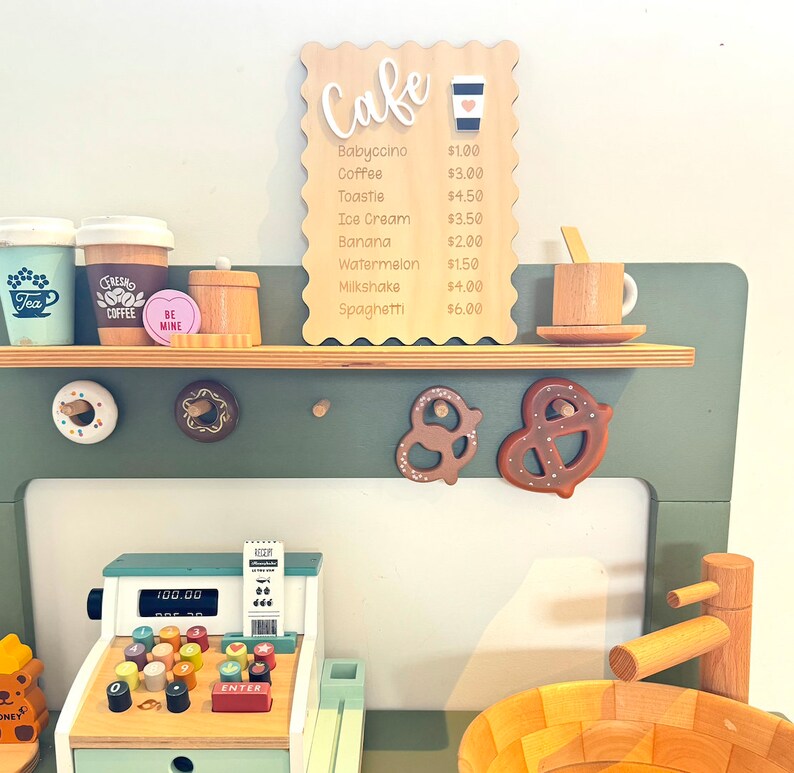 May include: A wooden toy cafe playset with a cash register, a wooden sink, and a menu board with prices for various food and drink items. The menu board says "Cafe" and lists items like "Babyccino", "Coffee", "Toastie", "Ice Cream", "Banana", "Watermelon", "Milkshake", and "Spaghetti".