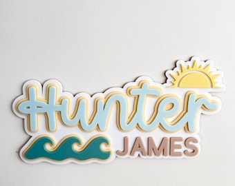 May include: A light blue and yellow layered name sign with the name "Hunter James" and a sun and waves design.