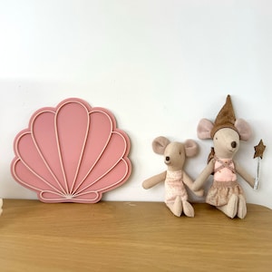 Peut inclure: Deux jouets en peluche en forme de souris, l'un portant un chapeau brun et une robe rose avec des accents dorés, l'autre portant une robe rose à pois blancs, assis devant un décor mural en forme de coquille rose.