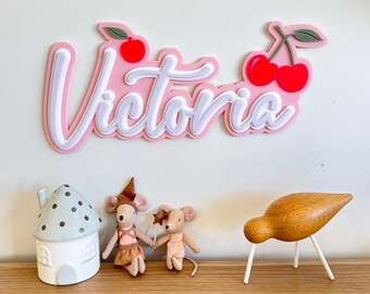 May include: A pink and white wooden sign with the name "Victoria" in white script with a pink outline. The sign is decorated with two red cherries with green stems and leaves.