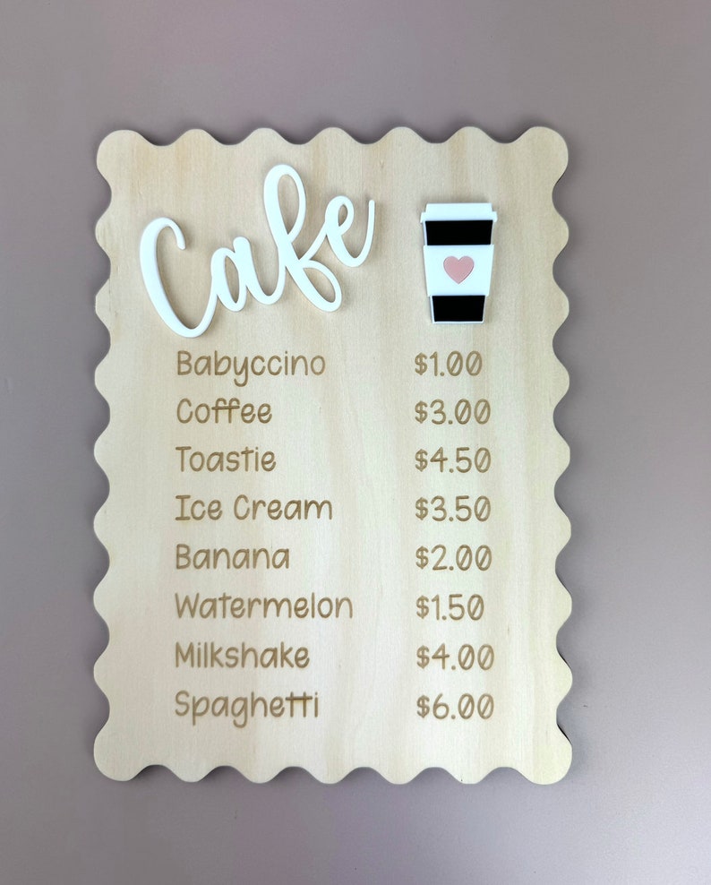 May include: A wooden sign with a wavy edge, painted in a light wood color. The sign says "Cafe" in white cursive letters with a small white coffee cup with a pink heart on it. The menu lists prices for items like babyccino, coffee, toastie, ice cream, banana, watermelon, milkshake, and spaghetti.
