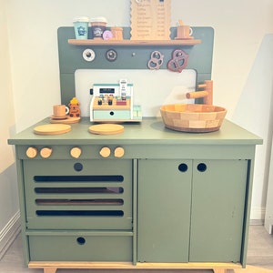 May include: A green wooden play kitchen with a stove, oven, and cabinets. The kitchen has a cash register, a sink, and a shelf with pretend food and drinks. The kitchen is designed for children to play pretend.