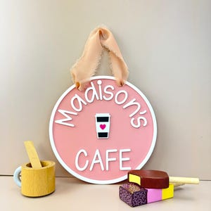 May include: A pink round sign with white trim that says "Madison's Cafe" with a coffee cup icon and a pink heart. The sign is hanging from a peach-coloured ribbon. There is a wooden toy cup and spoon and two wooden toy popsicles in the image.