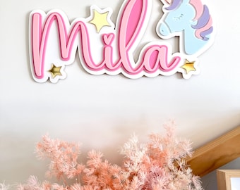 May include: Pink wooden name sign with the name "Mila" in a cursive font. The sign is decorated with gold stars and a blue unicorn head.