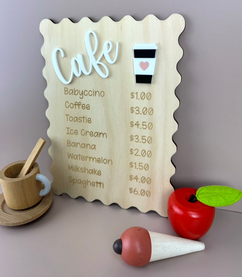 May include: A wooden sign with a cafe menu. The sign is painted brown and has a scalloped edge. The menu lists items like babyccino, coffee, toastie, ice cream, banana, watermelon, milkshake, spaghetti, and their prices. The sign is decorated with a small white coffee cup with a pink heart.