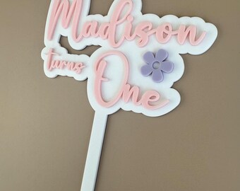 May include: Pink and white cake topper with the words "Madison turns One" and a purple flower.
