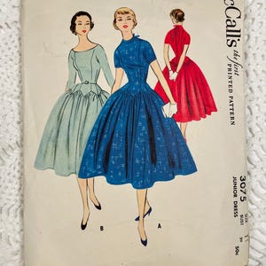 May include: A vintage McCall's printed pattern envelope for a junior dress, style 3075, size 11, bust 29. The illustration features three dresses: a light green dress, a blue patterned dress, and a red dress, all with full skirts and fitted bodices.