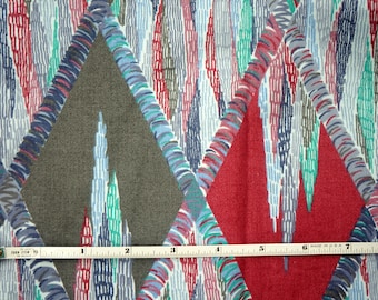 May include: Fabric with a repeating geometric pattern. The design features large diamond shapes in red, gray, and pink, outlined with blue and white. The background is filled with vertical lines in various colors. A measuring tape is across the fabric.