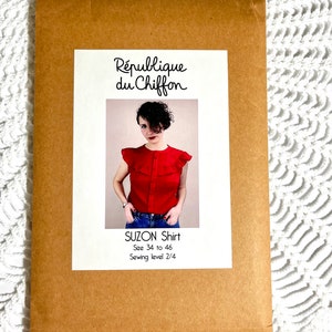 Puede incluir: Un sobre de papel marrón con una etiqueta blanca que dice "République du Chiffon" y una foto de una mujer con una camisa roja con cuello de volantes. La etiqueta también dice "SUZON Shirt, Talla 34 a 46, Nivel de costura 2/4".