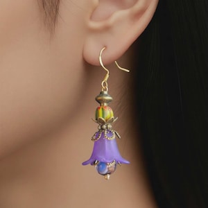 May include: A single gold-colored earring with a purple flower-shaped pendant. The pendant has a blue and gold bead at the bottom and a multi-colored bead above the flower. The earring is hanging from a gold-colored hook.