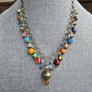 Può includere: Una collana in stile vintage con una catena in bronzo e una varietà di perline colorate. La collana presenta perline rotonde, sfaccettate e a goccia in tonalità di rosso, arancione, giallo, verde, blu e viola. Un grande ciondolo in bronzo pende al centro.