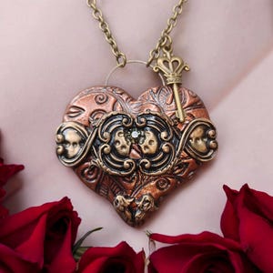 Könnte beinhalten: Eine herzförmige Halskette mit einem Schlüsselanhänger. Der Anhänger ist aus Kupfer und hat ein detailliertes Design mit einer schwarzen und goldenen Filigranarbeit. Der Schlüsselanhänger ist golden und hat eine Krone oben drauf.