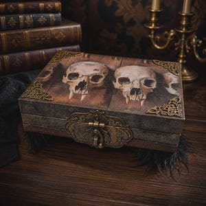 May include: A rectangular wooden box featuring a printed image of two human skulls on the lid. The box is accented with ornate, antique-style brass corner protectors and a central brass latch mechanism, providing a secure closure for the container.