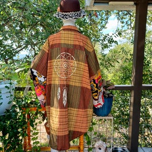 May include: A long, plaid shirt in shades of brown, orange, and yellow, featuring a white dreamcatcher design on the back. The sleeves have colorful floral and patterned cuffs. A brown hat with lace trim is on top.