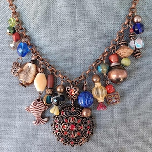 May include: A copper-toned necklace with a variety of colorful beads and charms. The necklace features a large, ornate pendant with red accents and a fish charm. Other charms include a heart, a blue sphere, and various glass beads.