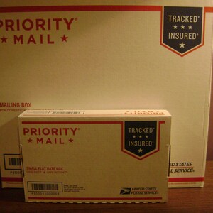 May include: Two Priority Mail boxes, one stacked on top of the other. The top box is a smaller flat rate box with the text "PRIORITY MAIL" and "TRACKED INSURED" in red and blue. The bottom box is a larger mailing box with the text "PRIORITY MAIL" and "TRACKED INSURED" in red and blue.