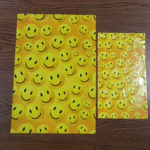 Puede incluir: Dos sobres de env&iacute;o de pl&aacute;stico amarillo con un patr&oacute;n repetitivo de caras sonrientes amarillas sobre una superficie de madera marr&oacute;n.