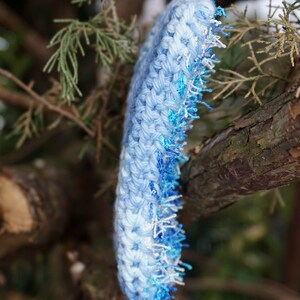 May include: A blue crocheted headband with a white and blue fringe. The headband is hanging from a tree branch.