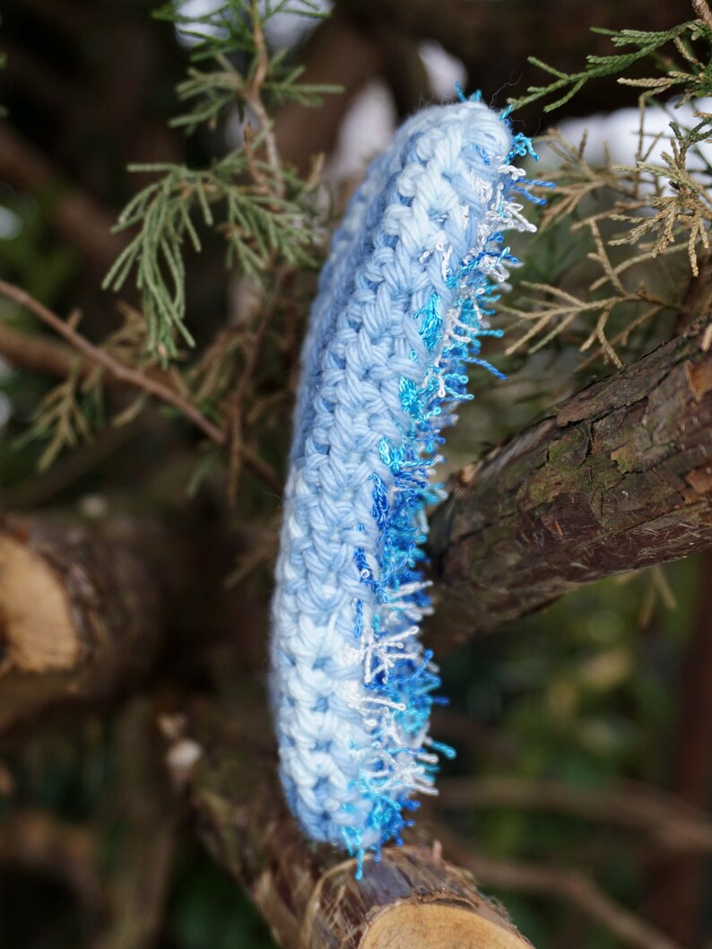 May include: A blue crocheted headband with a white and blue fringe. The headband is hanging from a tree branch.