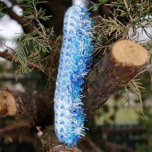 May include: A blue and white crocheted scarf with a fringe of blue and white yarn. The scarf is hanging on a tree branch.