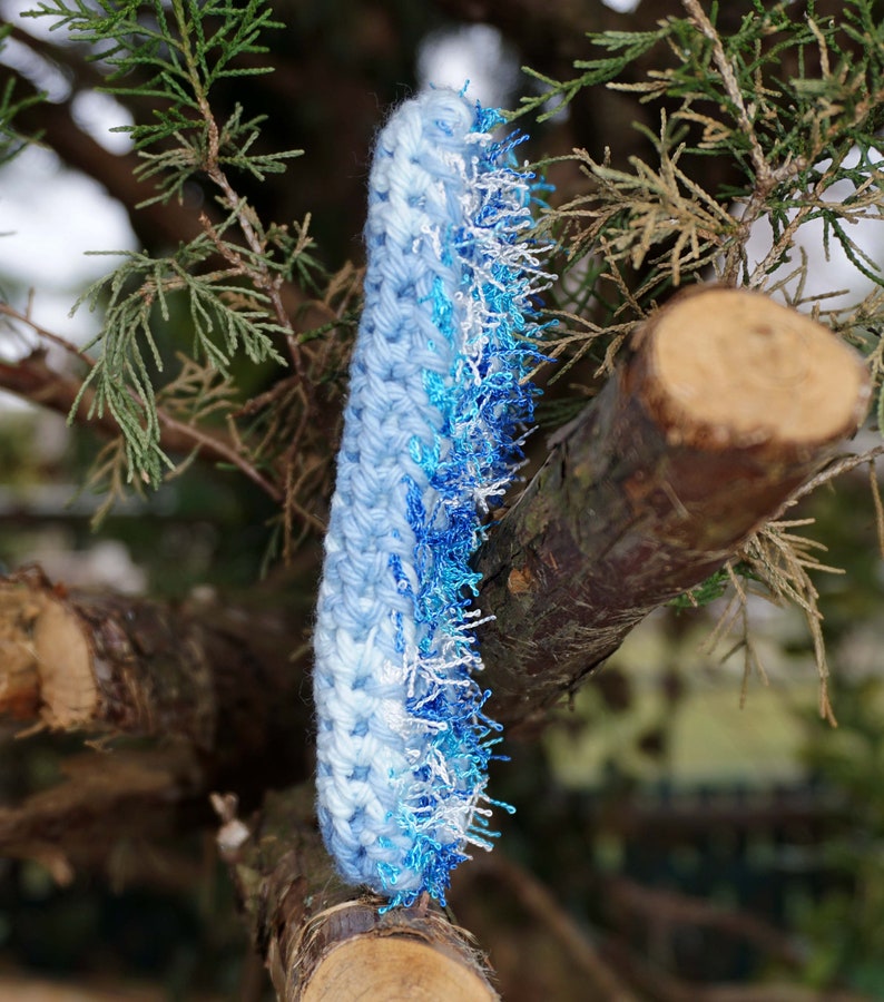 May include: A blue and white crocheted scarf with a fringe of blue and white yarn. The scarf is hanging on a tree branch.