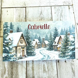 May include: A rectangular winter scene print featuring snow-covered houses and evergreen trees. The word "Gabrielle" is written in red script across the top. The artwork depicts a snowy village with a church and houses.