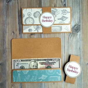 May include: A brown paper money holder with a nautical-themed design and a "Happy Birthday" message. The holder contains a fifty-dollar bill. The design includes a compass, shells, and a pocket watch.