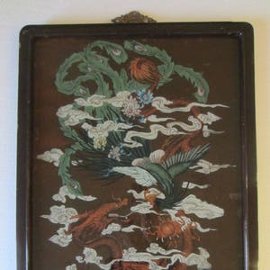 May include: A rectangular wall hanging featuring a dark brown background with a detailed illustration of a green phoenix and a red dragon intertwined among white stylized clouds. The artwork is encased in a dark brown frame with a metal hanger.