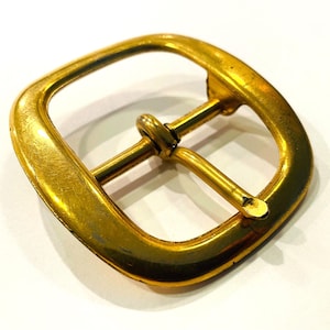 May include: A close-up of a gold-colored, square belt buckle. The buckle has a rectangular opening with rounded corners and a central bar for the belt. The metal has a slightly worn appearance.