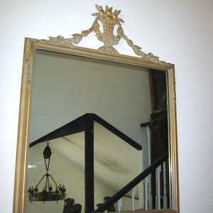 May include: A rectangular mirror with a gold-colored frame and ornate top detail. The mirror reflects a room with a chandelier, staircase, and dark beams. The frame has a decorative floral and basket design.