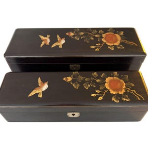 May include: Two rectangular black lacquer boxes featuring hand-painted gold and orange floral motifs and birds. Each box has a smooth finish and a small metal keyhole plate centered on the front edge, showcasing detailed botanical artwork.