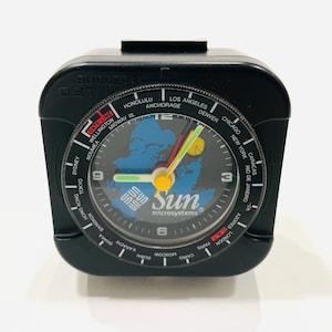 May include: Black travel alarm clock with a world map design. The clock face features city names and the "Sun microsystems" logo. The clock has a black exterior and a clear face.