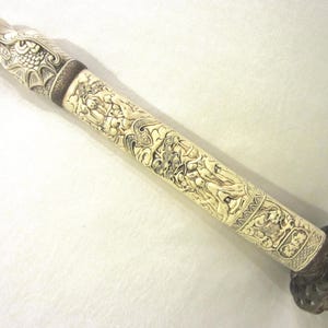 Puede incluir: Una empuñadura de espada con un mango cilíndrico tallado en color blanco hueso, con figuras en relieve intrincadas y un pomo en forma de dragón. La empuñadura incluye una guarda de metal oscuro y texturizado en la base con patrones detallados.