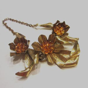 May include: A vintage gold-toned metal necklace featuring three dimensional flower and leaf designs. The flower centers are adorned with orange-colored rhinestones. The necklace has a delicate chain.