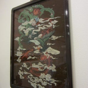 May include: A rectangular wall hanging featuring a dark brown background with a detailed illustration of a green phoenix and a red dragon intertwined among white clouds. The artwork is encased in a dark, polished wooden frame with a metal hanger.