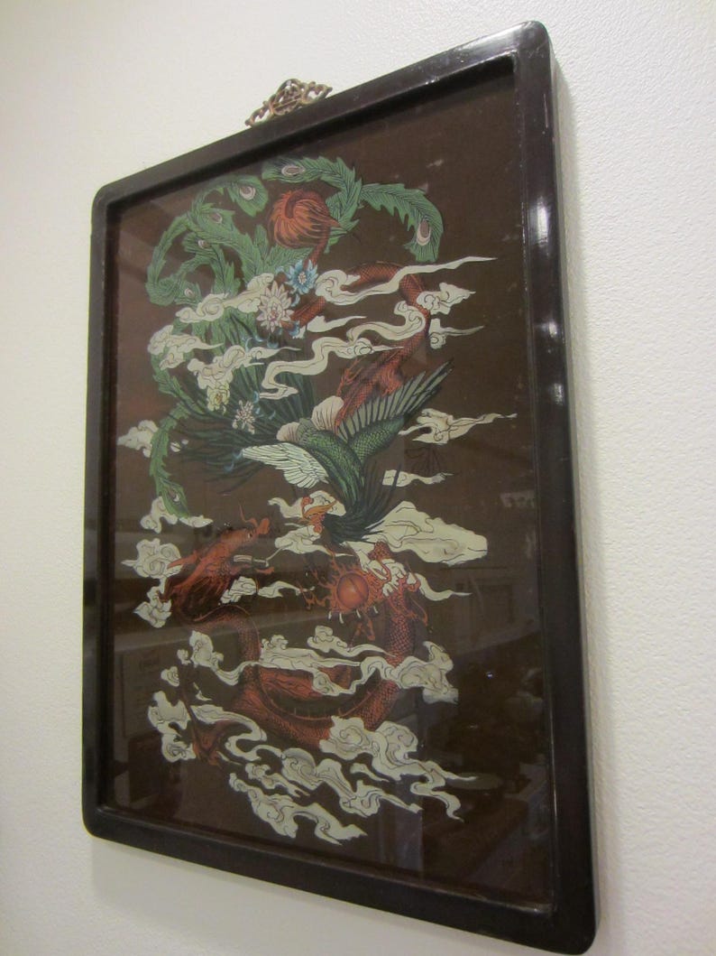 May include: A rectangular wall hanging featuring a dark brown background with a detailed illustration of a green phoenix and a red dragon intertwined among white clouds. The artwork is encased in a dark, polished wooden frame with a metal hanger.