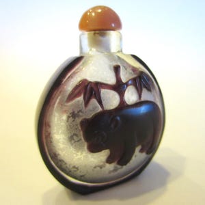 Puede incluir: Una pequeña botella de rapé de vidrio translúcido de forma redonda, con un relieve tallado en color púrpura oscuro que muestra un oso bajo una rama de bambú. La botella tiene un cuerpo liso y redondeado, rematado con un tapón esférico naranja claro.