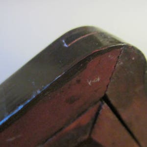 May include: A close-up view of a dark brown wooden corner joint with a smooth, polished finish. The structure features a curved edge meeting a flat side, revealing a reddish-brown interior tone where the wood grain is visible along the angled seam.