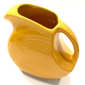 May include: A bright yellow ceramic pitcher featuring a smooth, glossy finish, a curved handle, and a wide pouring spout. The body of the pitcher displays subtle concentric ring patterns molded into the surface, creating a rounded silhouette.
