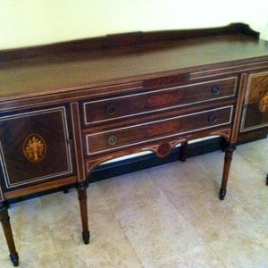 May include: A dark wood buffet with a rectangular top and two drawers. The buffet has decorative inlaid details and a cabinet door on each side. The legs are slender and tapered. The overall design is classic and elegant.