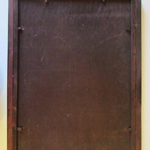 May include: A rectangular wooden frame with a dark brown finish and a flat, smooth backing panel. The frame features a small, ornate, metallic hanging bracket centered at the top edge, secured with two visible metal fasteners on the wood.