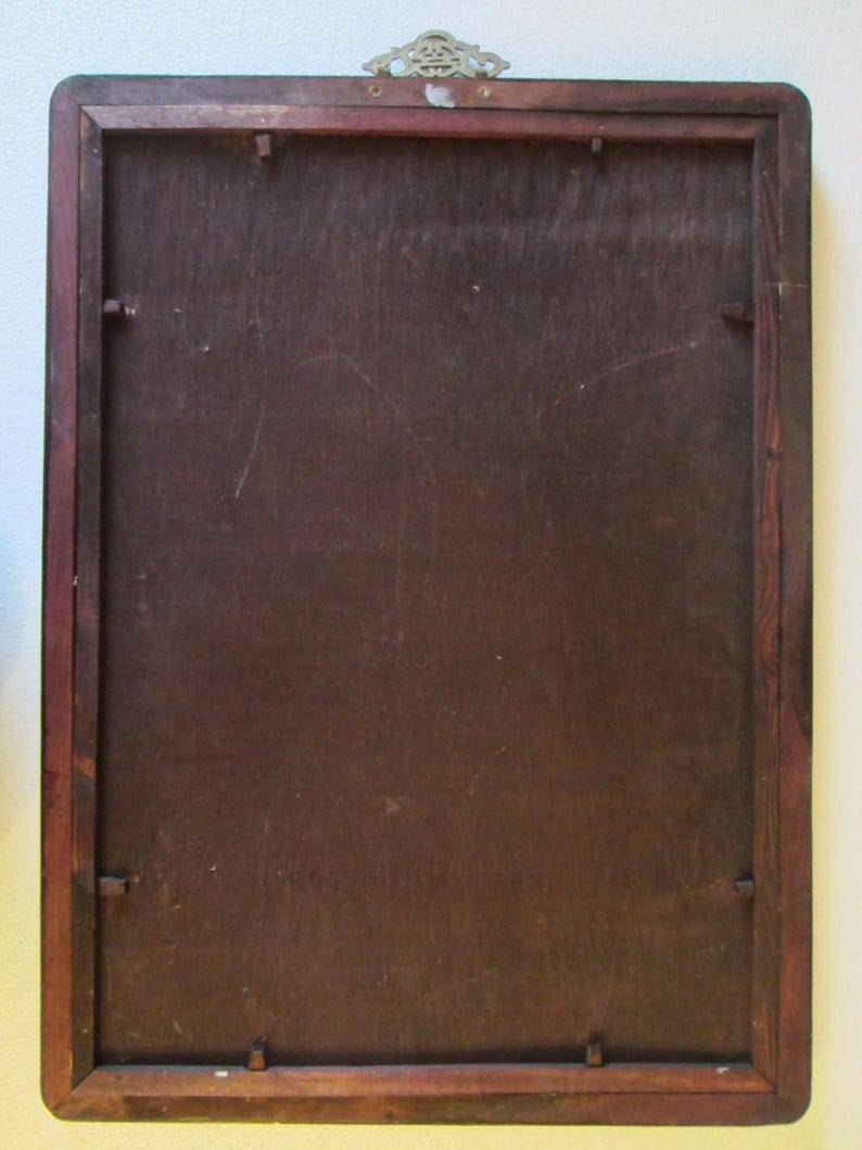 May include: A rectangular wooden frame with a dark brown finish and a flat, smooth backing panel. The frame features a small, ornate, metallic hanging bracket centered at the top edge, secured with two visible metal fasteners on the wood.