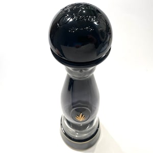 Peut inclure: Une grande bouteille en verre noir brillant avec un bouchon sphérique. Le sommet présente un texte en relief "Clase Azul" avec des ornements. Un petit emblème doré en forme de plante d'agave est centré sur le bas de la bouteille.