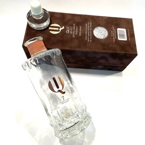 May include: A clear glass bottle of Qui Select Extra Añejo Tequila with a copper-colored neck band and a textured base. The bottle features the text Qui 7 Years Old 100% Agave. It is accompanied by a brown rectangular box with matching branding.