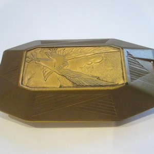 May include: A rectangular brass-toned metal case with beveled edges and geometric line patterns. The top surface features a raised relief design of a bird in flight with wings spread, clutching an object in its talons, set within a central panel.