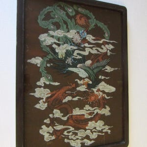 May include: A rectangular wall hanging featuring a dark brown frame and a glass panel. The artwork depicts a green phoenix and a red dragon intertwined amidst white stylized clouds on a dark brown background, with a metal hanger at the top.