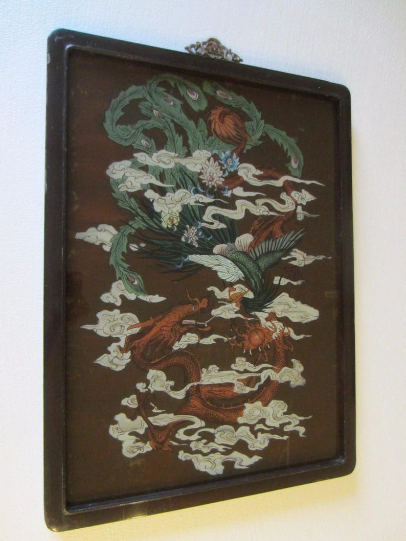 May include: A rectangular wall hanging featuring a dark brown frame and a glass panel. The artwork depicts a green phoenix and a red dragon intertwined amidst white stylized clouds on a dark brown background, with a metal hanger at the top.