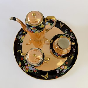 May include: A tea set featuring a teapot, a lidded sugar bowl, and a teacup with a saucer, all arranged on a round tray. The pieces have a peach-colored luster finish, black borders, and a colorful floral and butterfly pattern with gold trim.