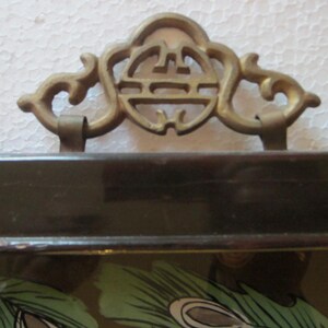 May include: A dark brown lacquered tray featuring a decorative brass handle with an intricate openwork design. The handle is attached to the top edge of the rectangular tray, which displays a partial green and black peacock feather pattern.